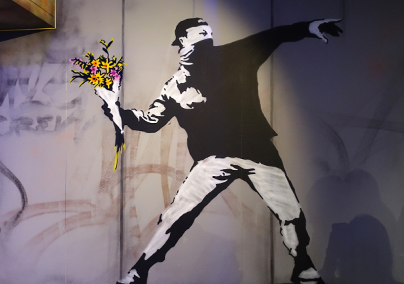 Opera di Banksy. Murales che ritrae un ragazzo mascherato che lancia un mazzo di fiori.