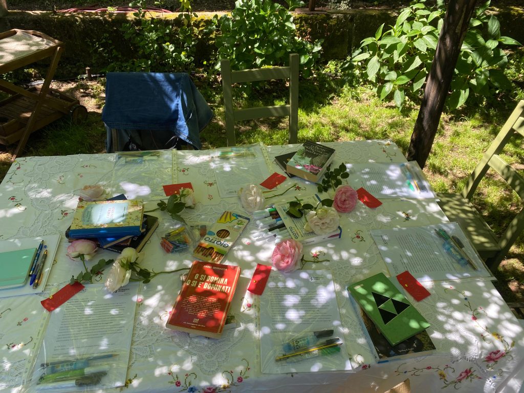 Lo spazio di lavoro creativo sotto un gazebo di piante e fiori. Sul tavolo una tovaglia di lino bianco, libri, fiori e colori