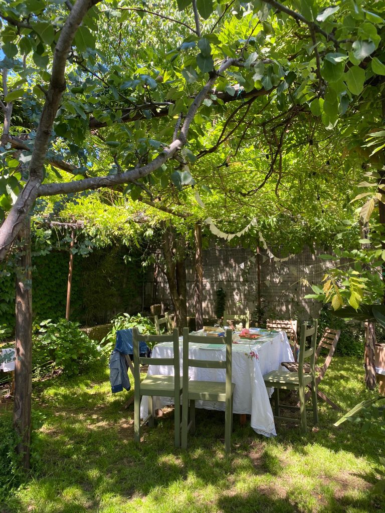 Lo spazio di lavoro creativo sotto un gazebo di piante e fiori. Sul tavolo una tovaglia di lino bianco, libri, fiori e colori