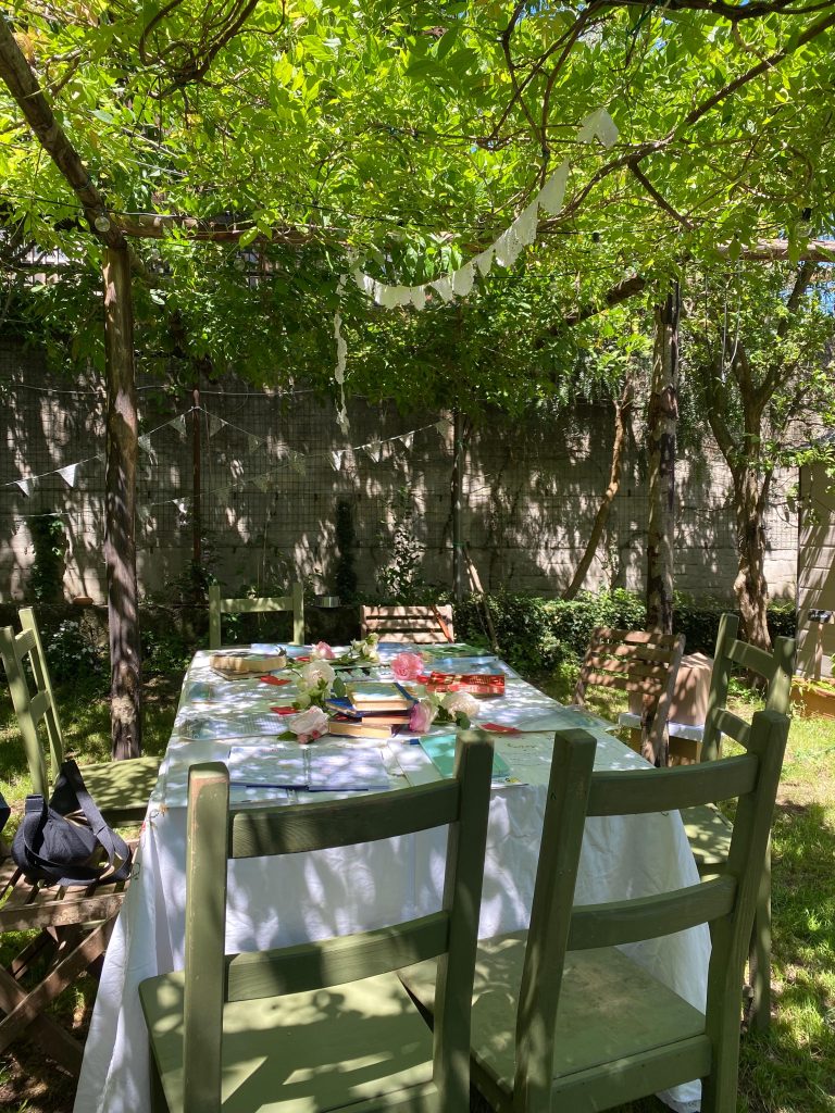 Lo spazio di lavoro creativo sotto un gazebo di piante e fiori. Sul tavolo una tovaglia di lino bianco, libri, fiori e colori